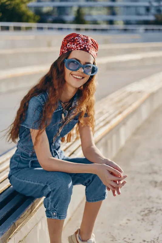 Lifestyle woman in modern boho-western denim street style wearing sunglasses and a red bandana, smiling outdoors in warm natural light with film color tones.