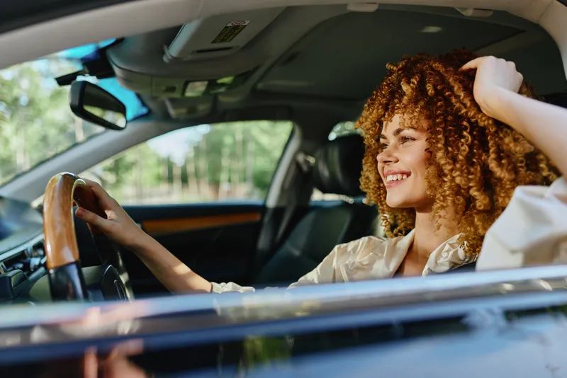 smiling woman driving car with curly hair, casual outfit, sunny day, joyful mood, natural light, green forest background