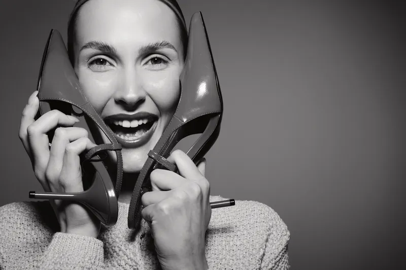 Smiling Caucasian white woman holds red high heel shoes near face, happy expression on black background, concept of fashion and footwear.