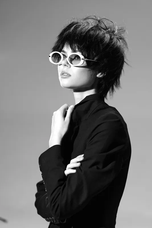 Black and white portrait of a stylish woman wearing retro sunglasses and dark clothing, expressing confidence and elegance.
