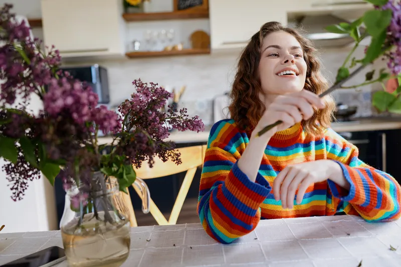 Young girl in a bright multicolor striped sweater relaxes at a sunlit kitchen table, smiling softly as fresh flowers sit nearby, creating a warm, cozy lifestyle moment