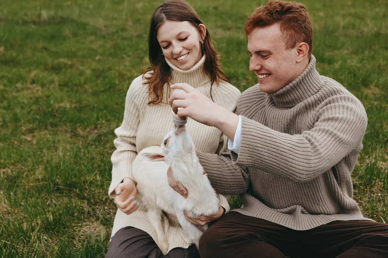Young couple in cozy sweaters enjoys feeding a white baby goat while sitting on grass in a relaxed outdoor lifestyle moment during autumn.