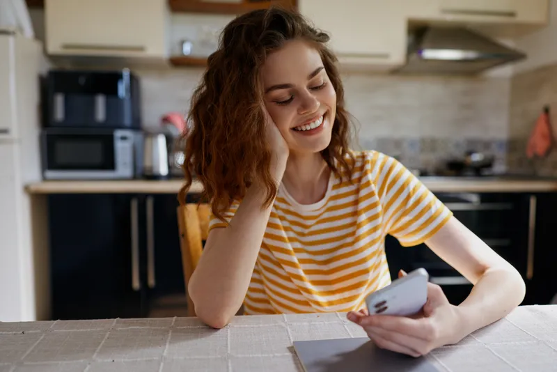 Young woman at home sits at a kitchen table, smiling while using a smartphone, capturing a casual lifestyle moment in a cozy domestic setting, bright natural light