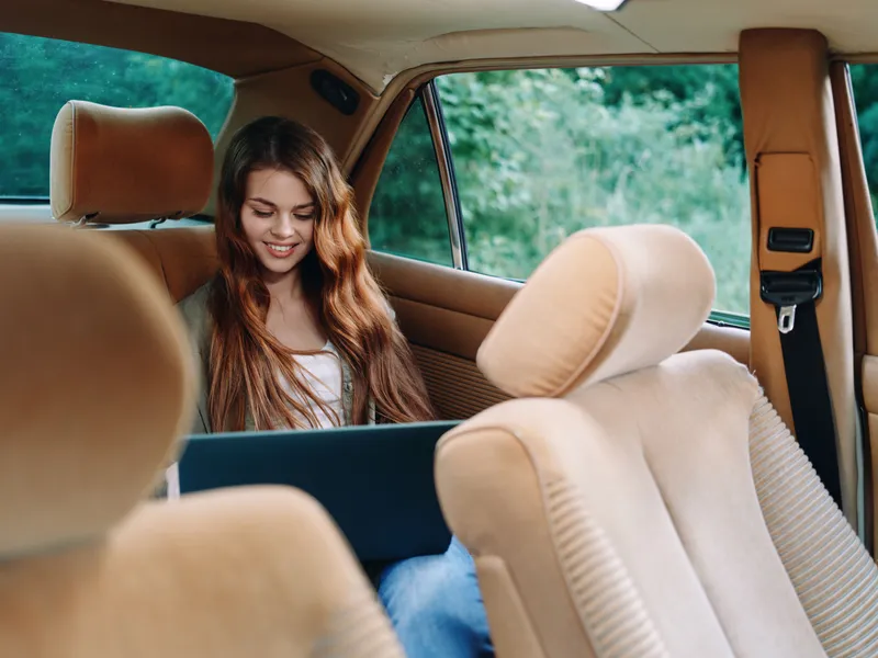 A cheerful young woman using a laptop in a cozy vintage car interior, surrounded by nature, reflecting a sense of freedom and creativity, perfect for lifestyle themes.