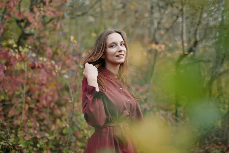 A candid portrait of a smiling woman in a burgundy dress standing among autumn foliage, conveying authentic natural presence, genuine demeanor, confident believable expression and relaxed posture in