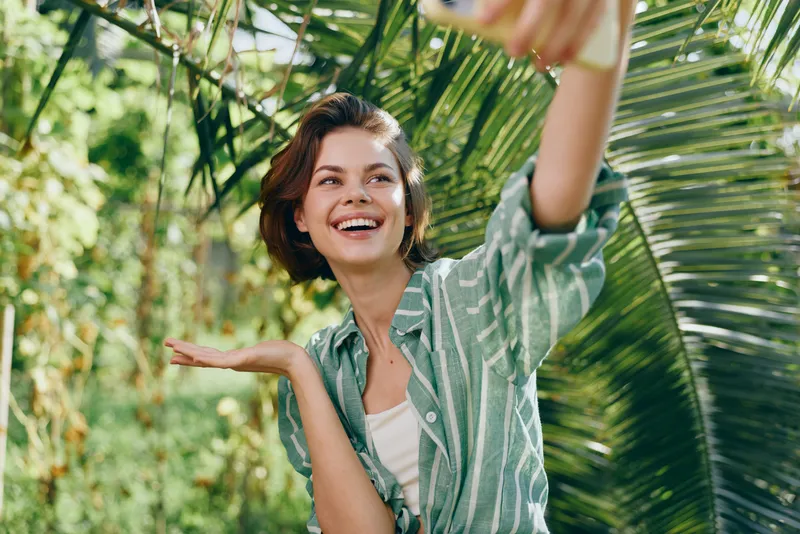 Woman, smiling, outdoor scene, cheerful pose among lush greenery, casual striped shirt, warm sunlight highlighting her joy, relaxed posture suggesting freedom and a fresh air moment