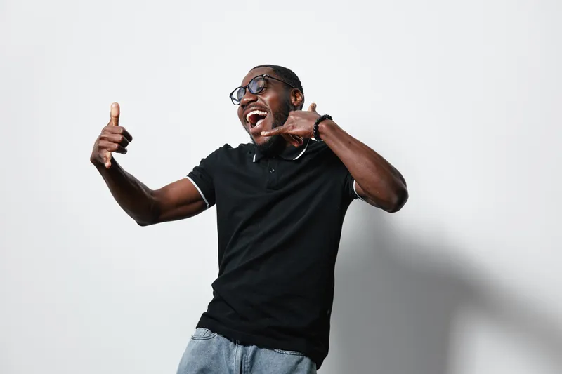 African American man with dark skin wearing glasses and black polo shirt making call me gesture and thumbs up with happy expression on white background.
