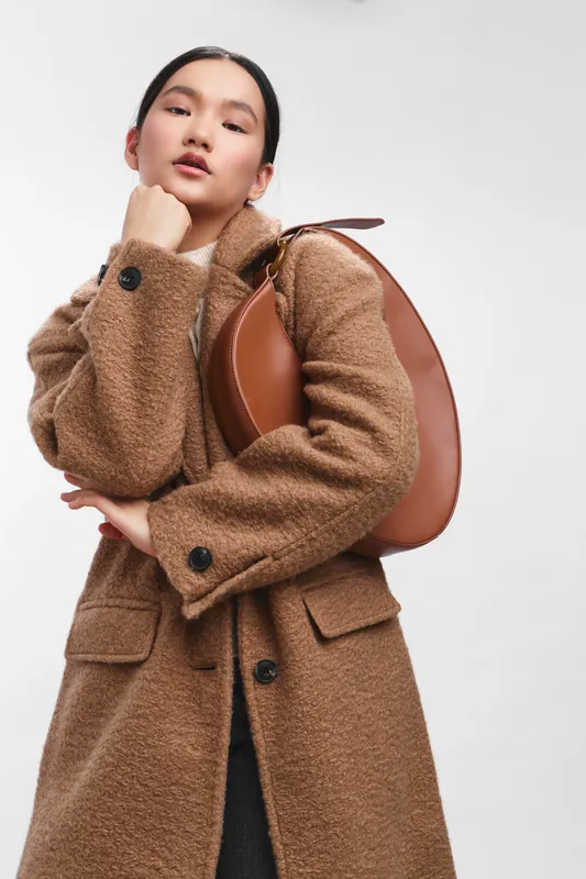 an asian woman stands in a studio wearing a brown wool coat, posing with a backpack, exuding confidence and contemporary fashion flair.