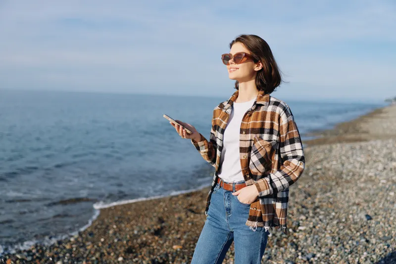 Woman on a beach with smartphone, casual plaid shirt, jeans and sunglasses, enjoying coastal view and fresh air along the shoreline under a clear sky.