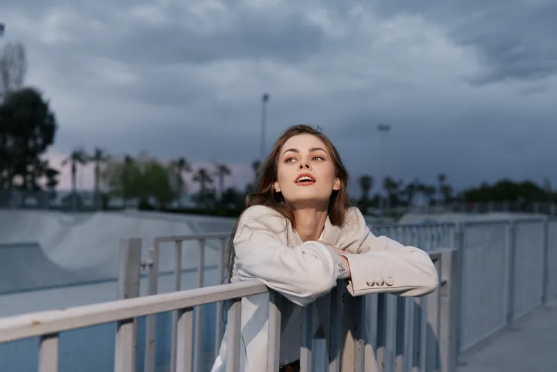 Businesswoman in light-colored blazer enjoying a moment of reflection outdoors, showcasing confidence and style against a dramatic sky background.