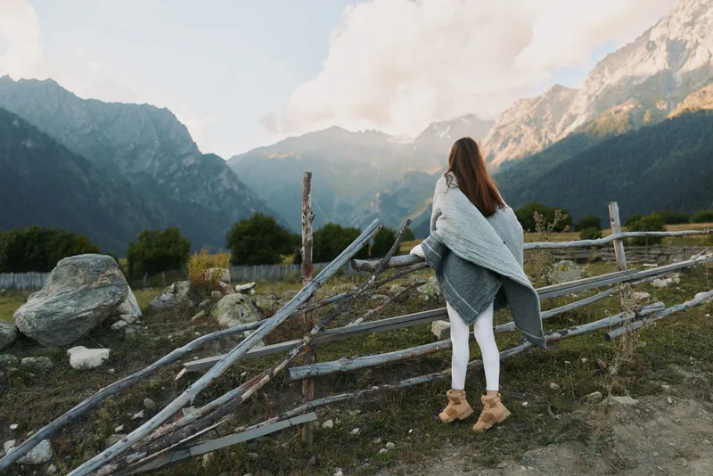 A woman stands in a rugged mountain landscape, wrapped in a cozy poncho, gazing toward distant peaks. A lifestyle moment highlighting outdoors, nature, and calm reflection.