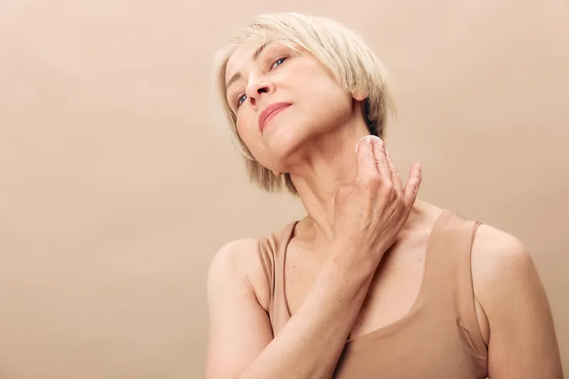 Confident mature woman with short blonde hair gently touching neck, wearing beige tank top. Soft neutral background highlights natural skin texture and graceful aging concept.