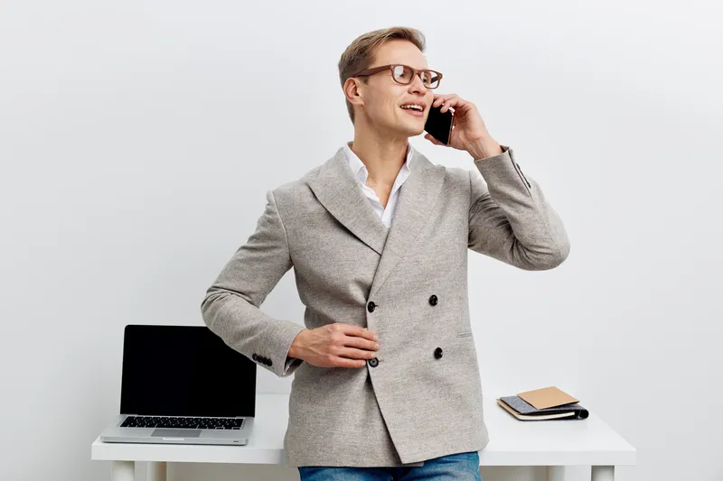Confident young man in stylish grey blazer and glasses smiling while talking on mobile phone in modern office background. Business professional lifestyle concept.