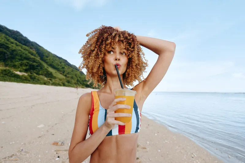 Woman with curly hair on sandy beach, holding juice cup with straw, smiling and relaxed, wearing swimsuit, ocean coastline behind, mountains in distance, sunny summer vacation mood
