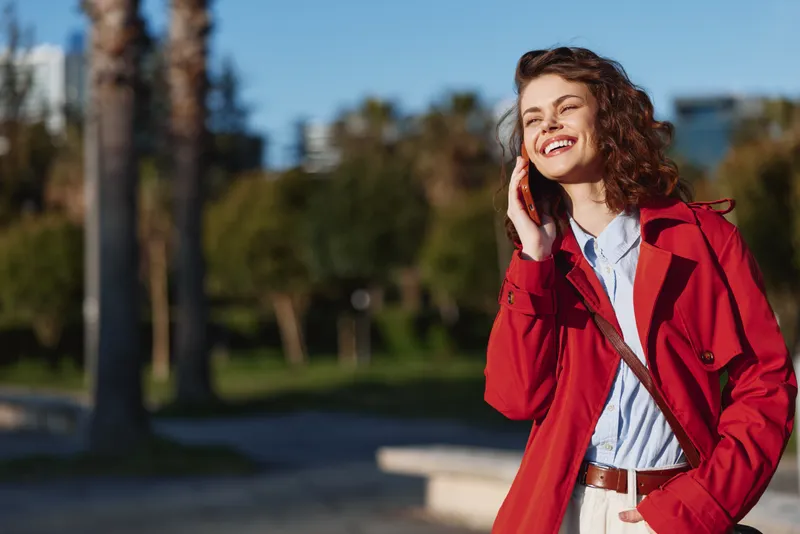 Young woman in red coat smiling talking on smartphone outdoors in park on sunny day, casual style, positive mood, urban background with trees and buildings.