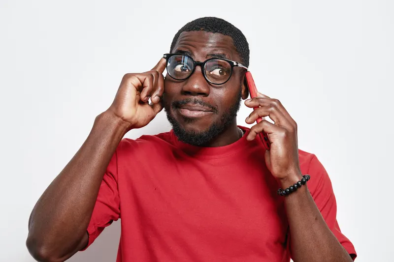 African American man with dark skin wearing glasses and red shirt talking on smartphone with thoughtful expression isolated on white background in studio.