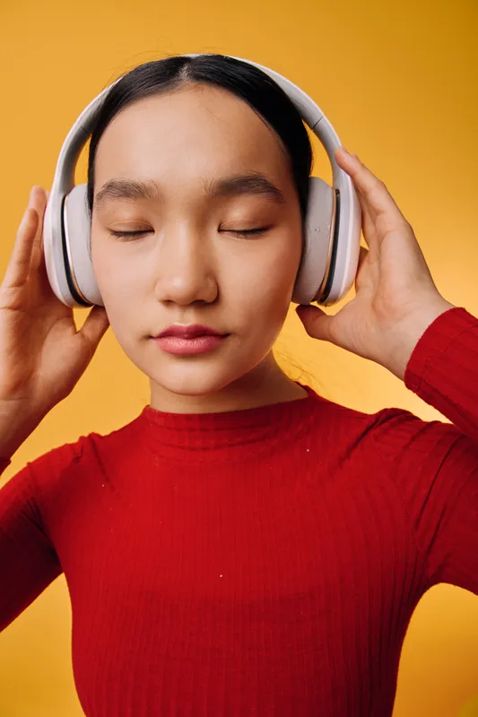Young woman with closed eyes wearing large headphones, enjoying music or meditation in a casual red sweater against bright yellow background in studio portrait.
