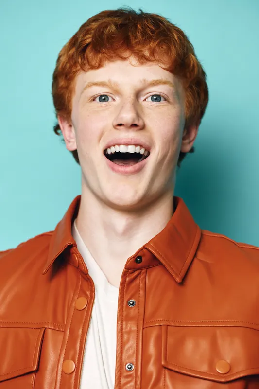 Young man with red hair wearing orange leather jacket smiling and looking excited in studio with turquoise background. Portrait of happy male model with open mouth and casual style.