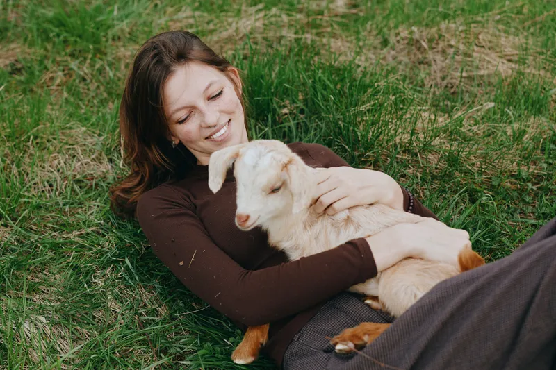 A young woman in a brown sweater lies on grass smiling while gently holding a white baby goat in a peaceful outdoor lifestyle scene.