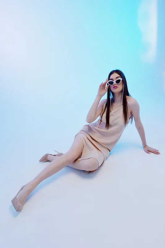 Young woman beauty fashion model wearing beige dress and sunglasses sitting on floor in studio with blue background. Concept for style, elegance, and modern trends.