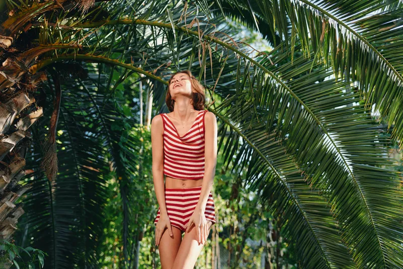 A cheerful traveler stands among lush palm fronds wearing a red striped bikini, smiling toward the sun, radiating carefree vacation vibes and tropical warmth.