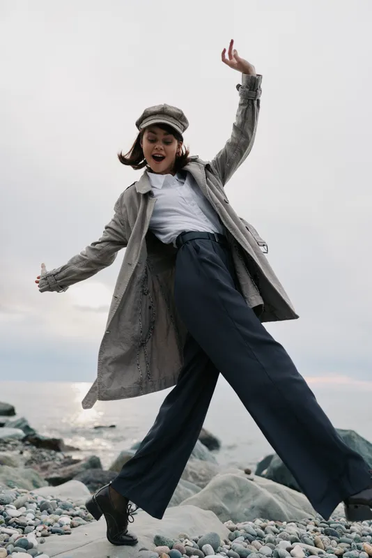 Young woman wearing a long coat and hat joyfully jumps on rocky beach near sea under cloudy sky. Casual fashion, outdoor, freedom, and happiness concept with natural light and dynamic pose.
