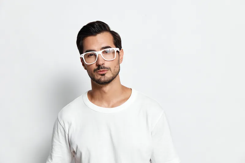 Stylish confident young man wearing white shirt and glasses looking at the camera with a serious expression against a plain solid background. Fashion concept.