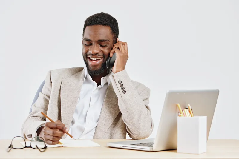Smiling man talking on mobile phone while working at desk in bright modern office, using laptop, notes and stationery, professional casual attire. People lifestyle concept, office communication, remot