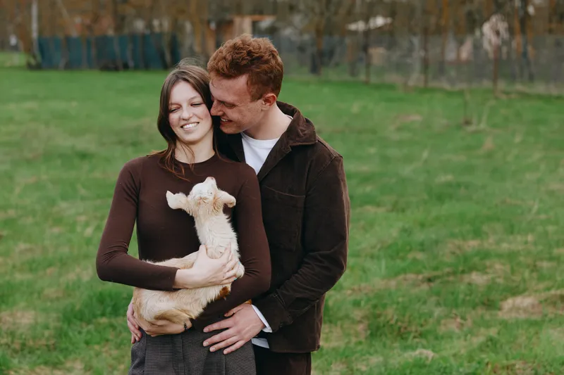 A joyful couple shares a moment with a small goat kid in an open grassy park, embracing and smiling. A warm lifestyle scene that captures affection, companionship, and simple countryside moments.