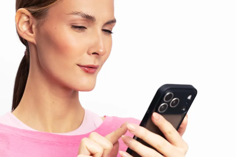 Young woman in pink sweater uses smartphone, focused on screen. Isolated on white background. Concept of technology, communication, and mobile device usage.