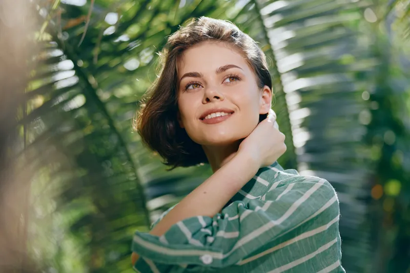 Happy young woman with short dark hair stands among lush greenery, wearing a striped green shirt, catching soft sunlight.