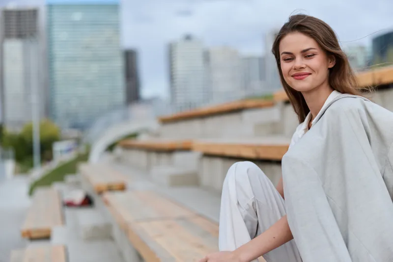 Woman in casual light clothing sitting on urban wooden bleachers with modern cityscape background peaceful smile autumn season lifestyle fashion