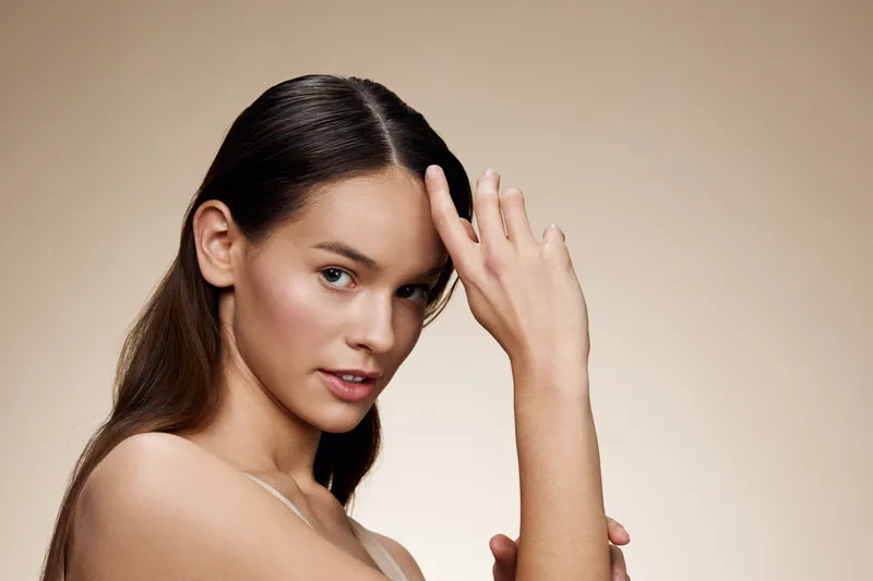 Young woman with long hair poses elegantly, showcasing beauty and confidence against a soft beige background, perfect for beauty and health themes