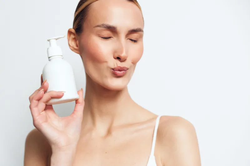 Young woman with clear skin holding white cosmetic bottle, eyes closed, making playful face, skincare concept, minimal background, natural light, beauty routine.
