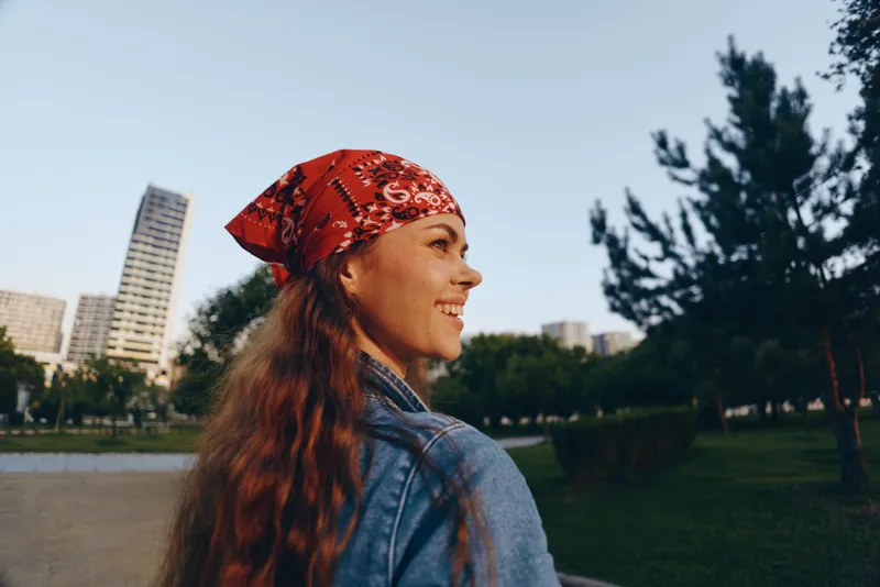 smiling woman wearing red bandana and denim jacket enjoying sunny day in urban park with green trees and modern buildings in background