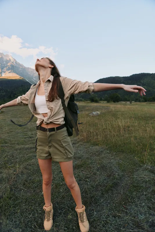 Young woman with long brown hair enjoys freedom with arms outstretched in a mountain meadow wearing khaki shorts, beige boots, and a tied shirt under clear blue sky.