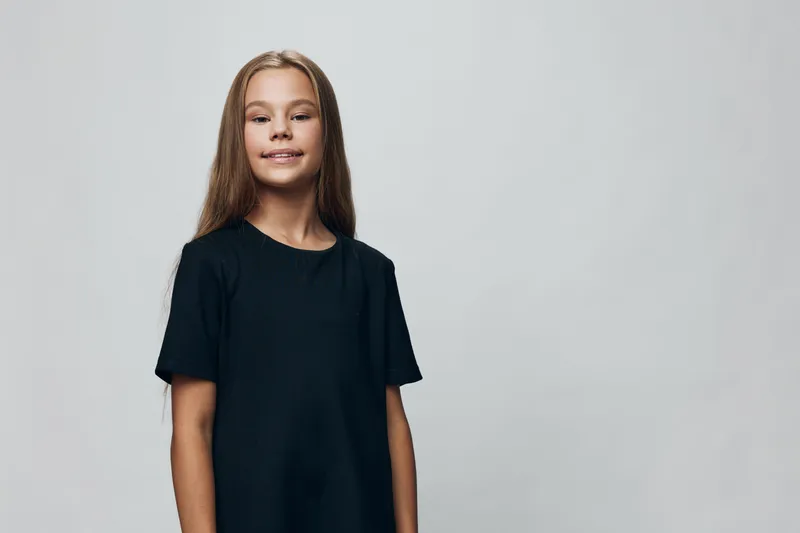 Girl child with long hair wearing black t shirt standing with smile and confidence on grey background. Portrait of happy preteen female with casual outfit in studio lighting.