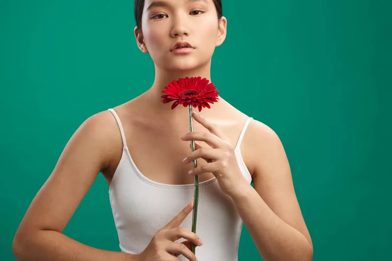 an asian woman stands against a green backdrop, holding a red flower near her collarbone with a calm expression. soft studio lighting highlights smooth skin and minimalist attire, creating a serene,