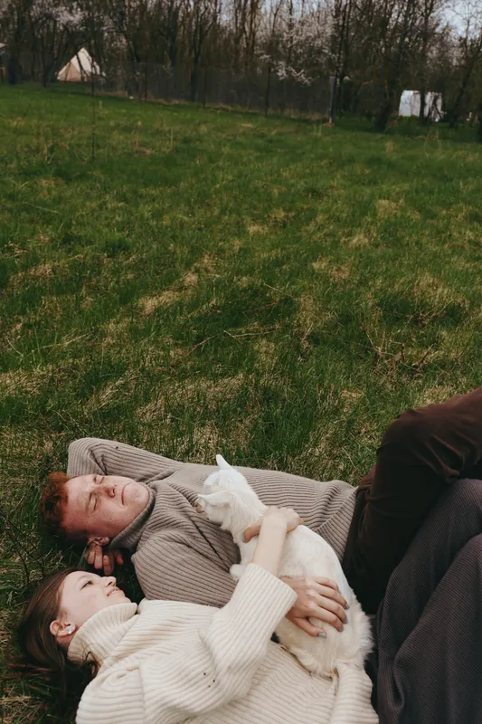 A relaxed lifestyle scene with two adults lying on a grassy field, cuddling a small white goat kid. Natural light, casual clothing, and a tranquil outdoor moment convey comfort, affection, and simple