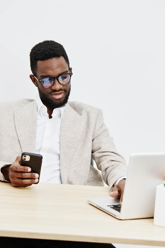 Professional young black man wearing glasses and formal jacket using smartphone while working on laptop at desk, isolated on plain white background, studio portrait. Business and technology concept