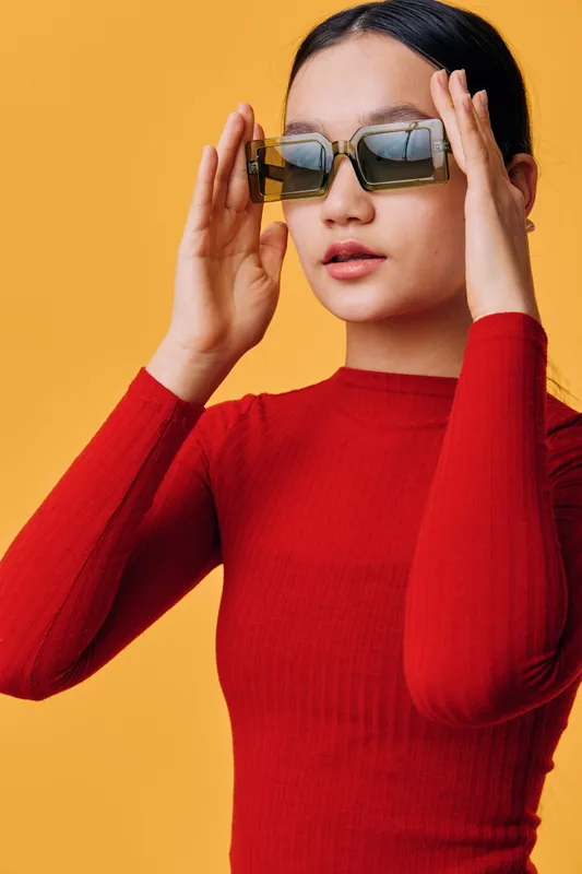Woman in red sweater wearing fashionable sunglasses with short black hair posing on yellow background. Portrait of trendy female model adjusting eyewear for style and confidence.