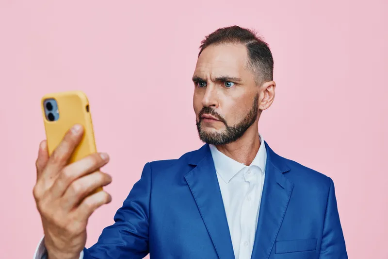 A professional man in a blue suit appears focused and puzzled as he photographs himself against a solid pink background, creating a modern business portrait.