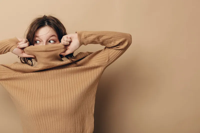 Young woman in brown knitted sweater pulls collar over face, looking sideways with curious expression. Neutral beige background creates warm, cozy atmosphere and casual style concept.