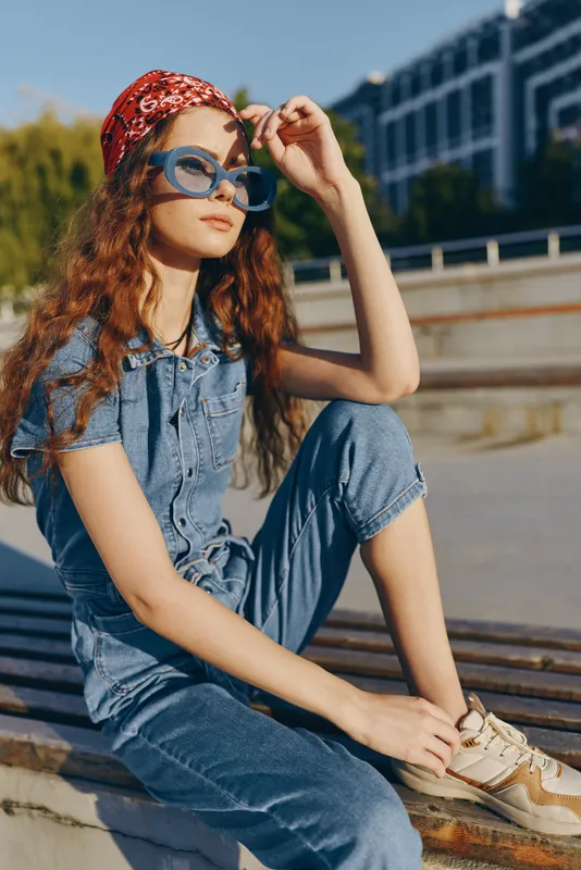 Lifestyle woman in modern boho-western denim street style wearing blue sunglasses and a red headscarf, sitting outdoors in warm sunlight, film color effect.
