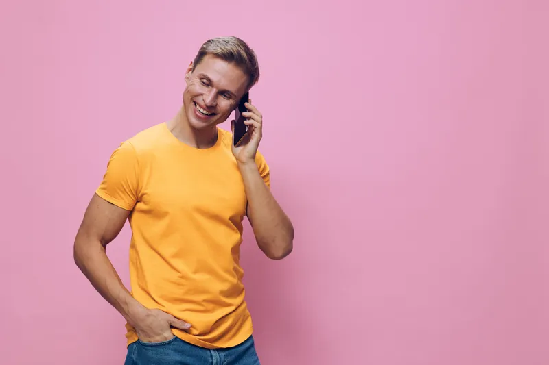 Smiling young man wearing yellow t-shirt talking on smartphone looking down isolated on pink background digital nomad lifestyle cheerful emotion concept