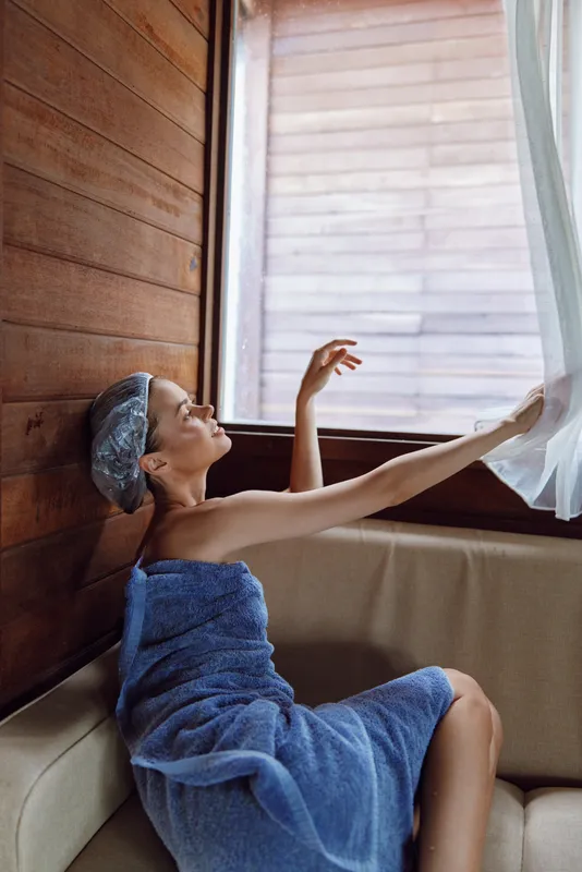 woman wrapped in blue towel and shower cap relaxing on beige sofa by wooden walls and soft sunlight through sheer curtains in cozy room interior