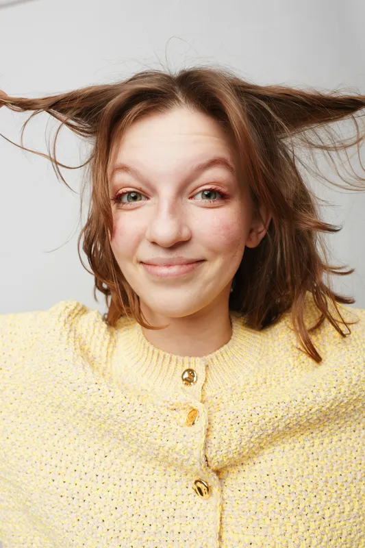 Smiling young woman with messy hair wearing a yellow knitted sweater looks directly at the camera. Studio portrait on a light background with playful and casual mood.