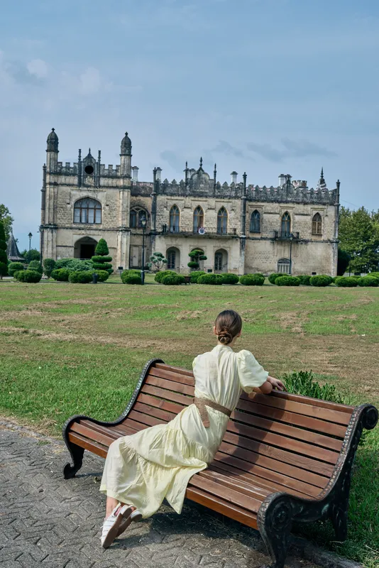 Woman in a vintage dress sits on a wooden bench in a park, facing a historic castle, creating a serene nostalgic scene with timeless architecture and lush surroundings.