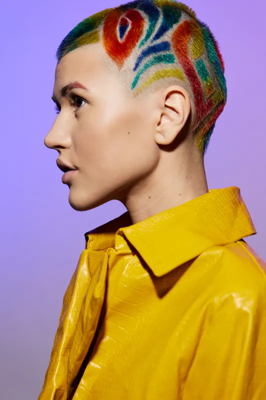 Woman with colorful shaved hair in a yellow jacket, showing artistic style and unique fashion against a soft purple background, capturing modern creativity and boldness.