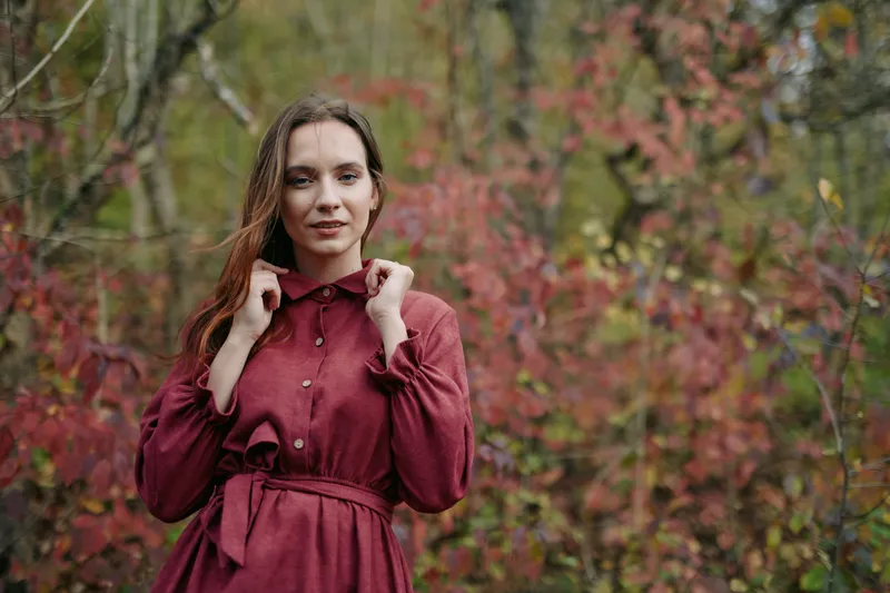 Portrait of a young woman in a burgundy dress standing in an autumn forest, conveying authenticity and believability through natural light and a relaxed, candid pose.